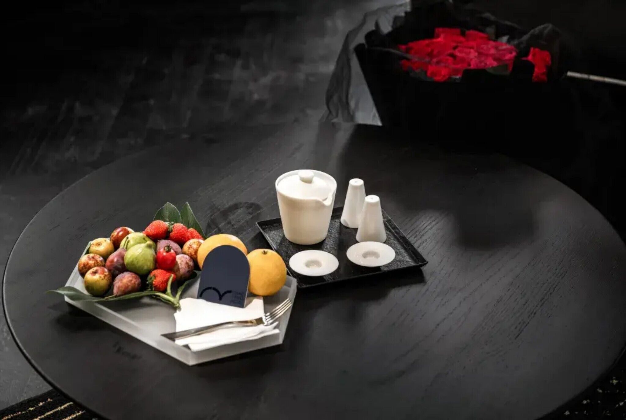 A black table with a white teapot, sugar cubes, a small plate of fruit, and a napkin with a fork.