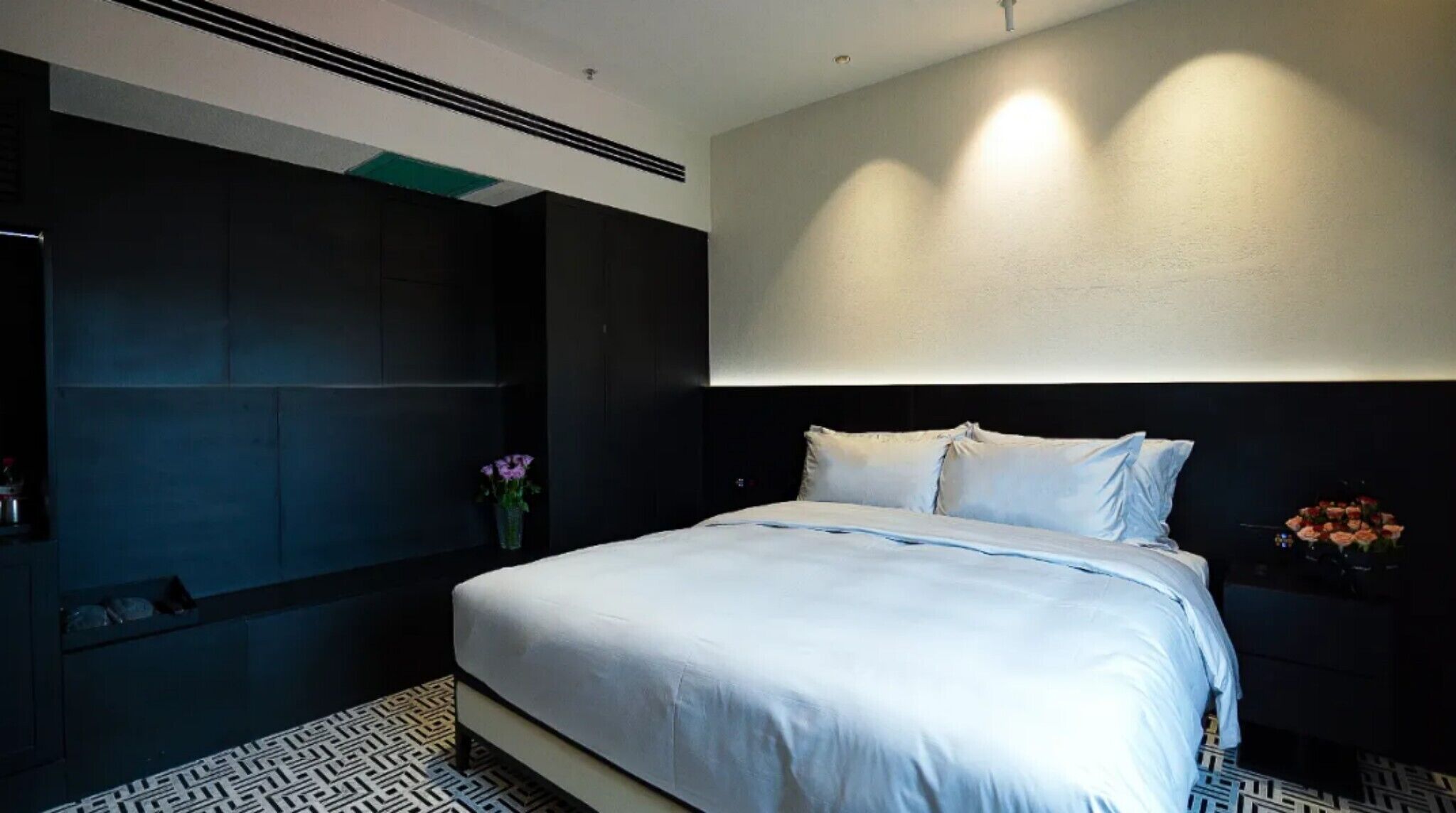 A modern bedroom with a large bed, a dark wall, and a small bedside table with a vase of flowers.