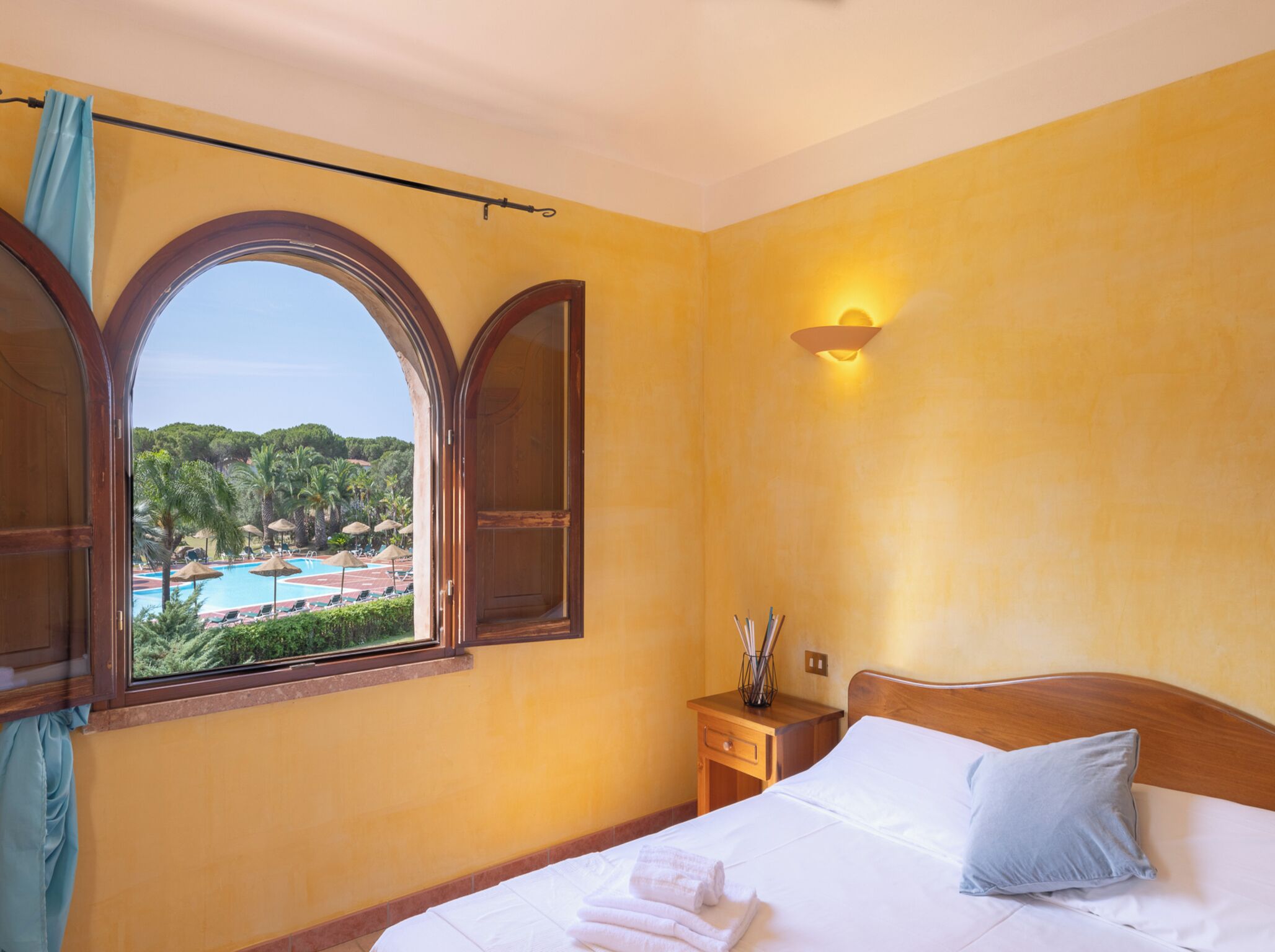 Ein Hotelzimmer mit Bett, Nachttisch, Fenster mit Blick auf Pool und Palmen sowie einer Wandleuchte.
