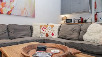 A living room with a grey sofa, a wooden coffee table with a book and a coaster, a colorful abstract painting, a kitchen area with cabinets and appliances, and a fluffy white throw pillow.
