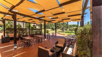 Pátio externo com móveis de vime e pérgola de madeira que oferece sombra.