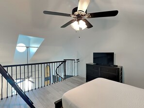 A modern bedroom with a ceiling fan, a TV, a bed, and a staircase leading to an upper level. - Treasure Cove #D308 | Lagoon Views, Near Beach + Amenities (Panama City Beach)