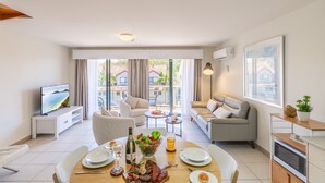 A modern living room with a dining area, a sofa, a TV, and a balcony with a view of buildings. - Bay Breeze Apartments, 33   1 Trafalgar St - Aircon, Wi-Fi, Linen, Pool (Nelson Bay)
