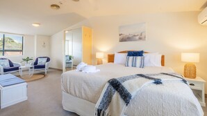 A hotel room with a large bed, a sitting area with a chair and a small table, a mirror, and a painting on the wall. - Bay Breeze Apartments, 33   1 Trafalgar St - Aircon, Wi-Fi, Linen, Pool (Nelson Bay)