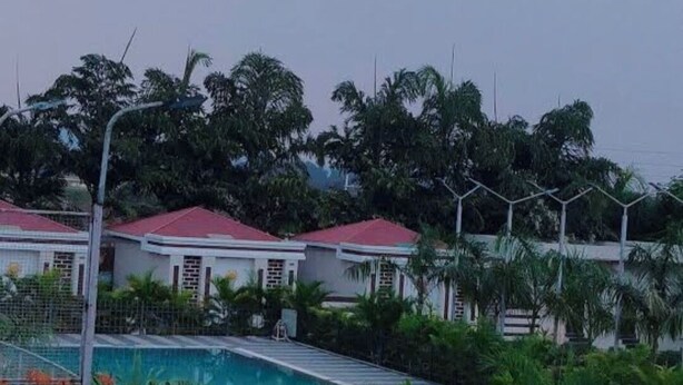 A pool area with a clear blue pool, surrounded by lush greenery and a few buildings with red roofs. - utsav hotel and resorts (Burhanpur)