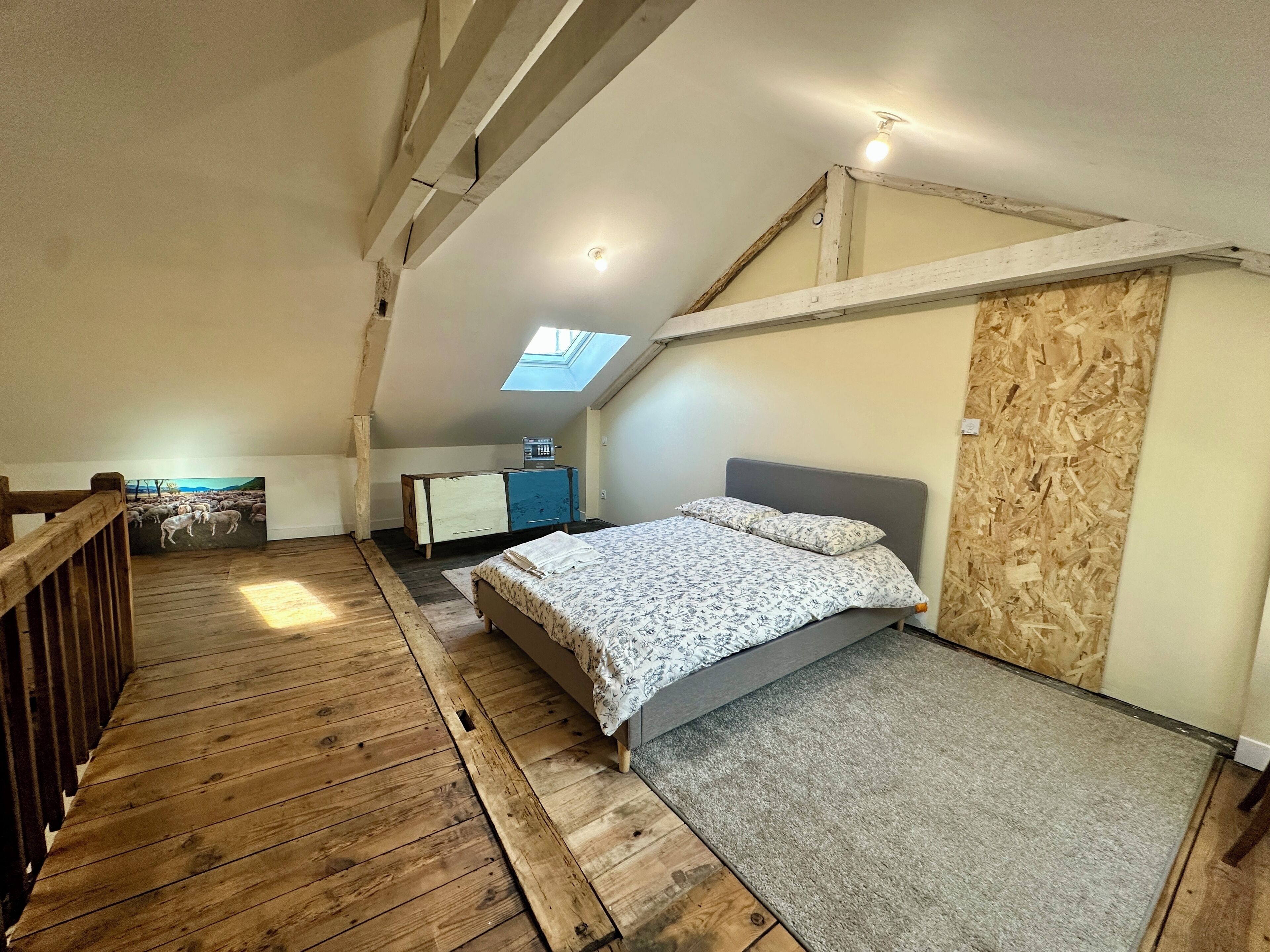 Ein Schlafzimmer im Spitzboden mit schrägem Dach, Holzboden, einem Bett mit grauem Kopfteil, einem kleinen Schreibtisch und einem Dachfenster.