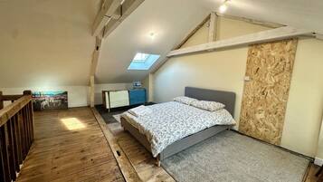 Ein Schlafzimmer im Spitzboden mit schrägem Dach, Holzboden, einem Bett mit grauem Kopfteil, einem kleinen Schreibtisch und einem Dachfenster.