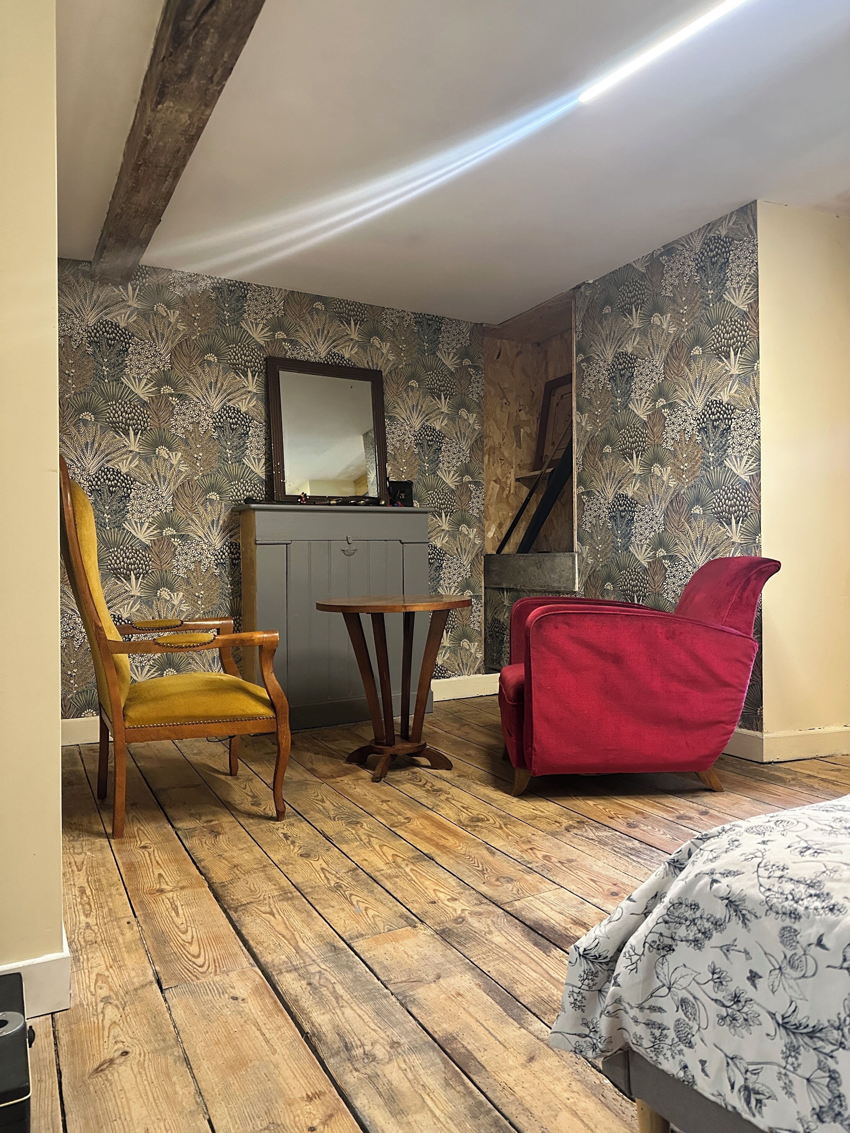 A room with wooden flooring, a wooden chair, a small round table, a red chair, and a bed with a floral-patterned bedspread.