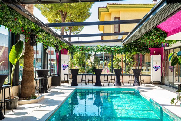 Ein Poolbereich mit klarem, blauem Pool, umgeben von Tischen und Stühlen, und einem überdachten Bereich mit einer Pergola.