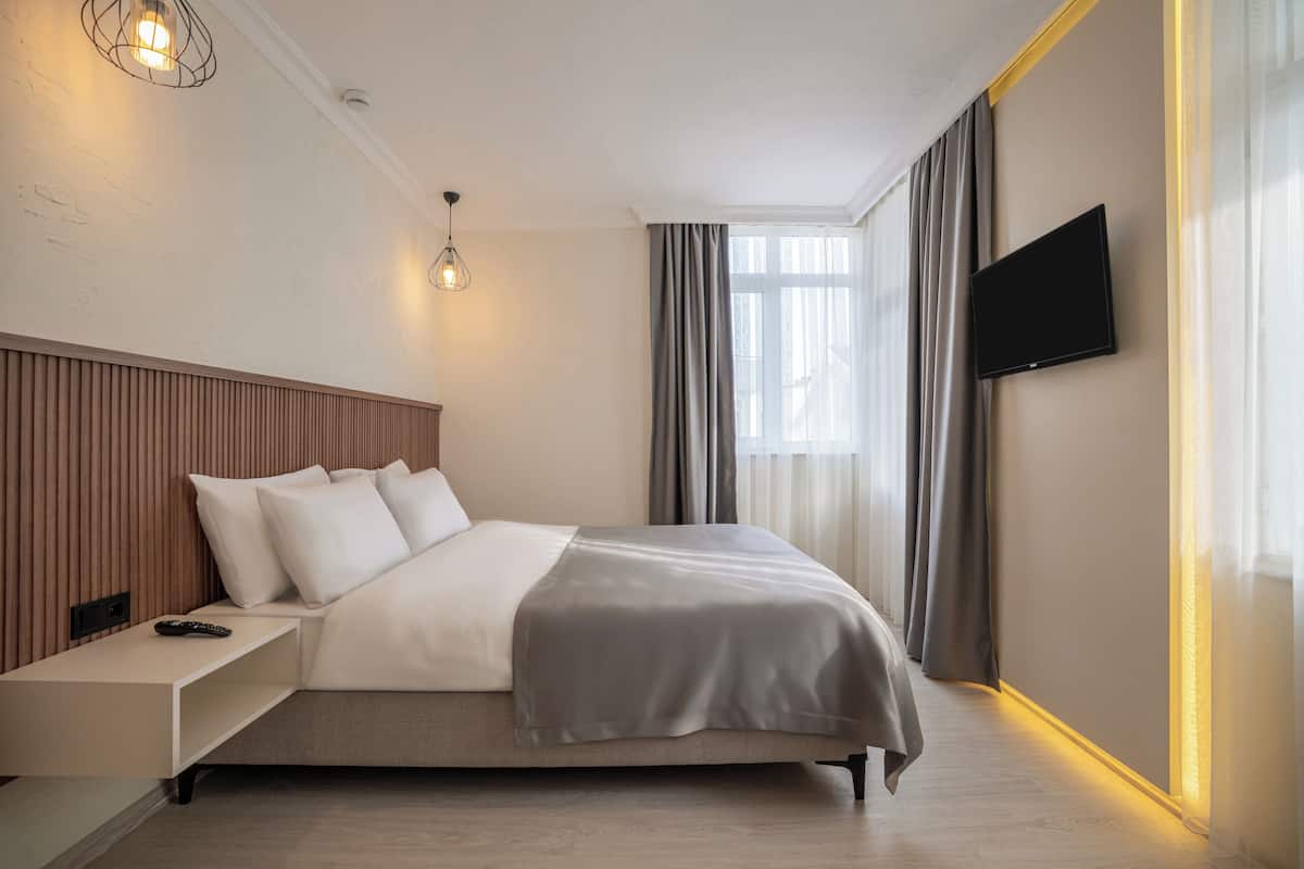 A modern hotel room with a large bed, a bedside table with a remote control, a wall-mounted TV, and a window with curtains.