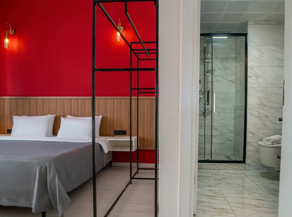 Ein modernes Hotelzimmer mit einer roten Akzentwand, einem Bett mit weißer Bettwäsche, einem Nachttisch und einer gläsernen Duschkabine.