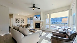 A spacious living room with a fireplace, a large window with a view, a ceiling fan, and a television.