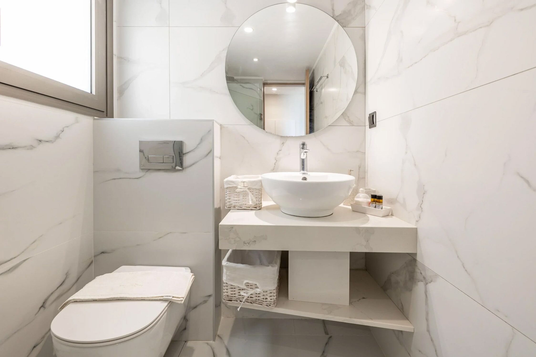 A modern bathroom with a white marble sink, a round mirror, and a toilet with a towel.