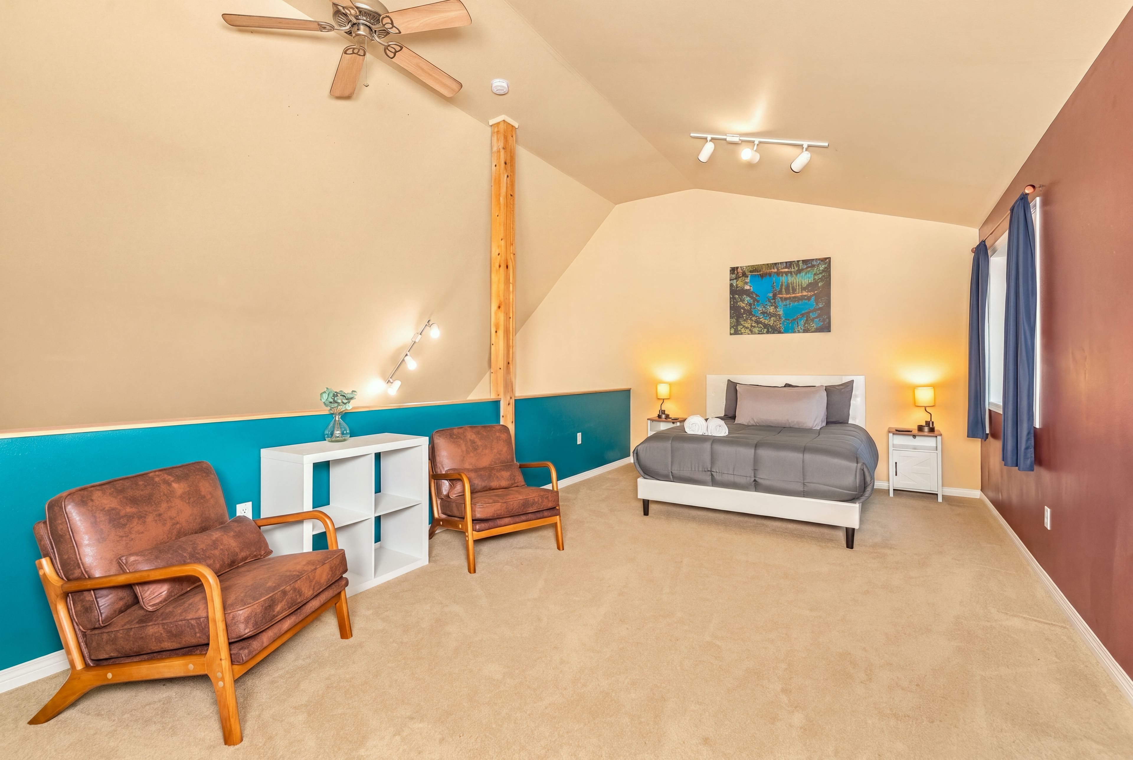 A bedroom with a bed, two chairs, a nightstand, a ceiling fan, and a painting on the wall.