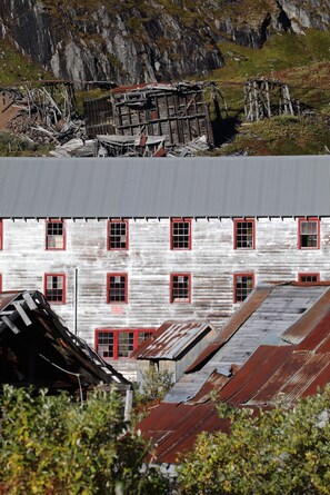 Un edificio rústico con varias ventanas y techo de metal, rodeado de terreno rocoso y vegetación.