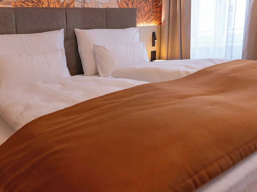 Un lit bien fait, avec du linge de lit blanc et un plaid marron, une tête de lit au motif texturé et un luminaire fixé au mur.