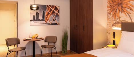 A hotel room with a bed, a dining table with chairs, a painting of a cityscape, and a large plant.