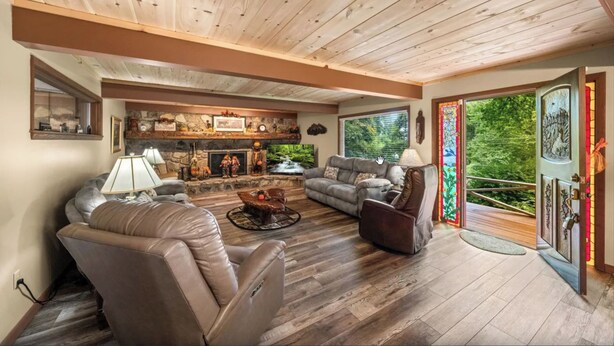 A spacious living room with leather sofas, a fireplace, and a view of a forest through large windows. - Gatlinburg Cabin with Hot Tub, Game Room, Sleeps 12 (Gatlinburg)