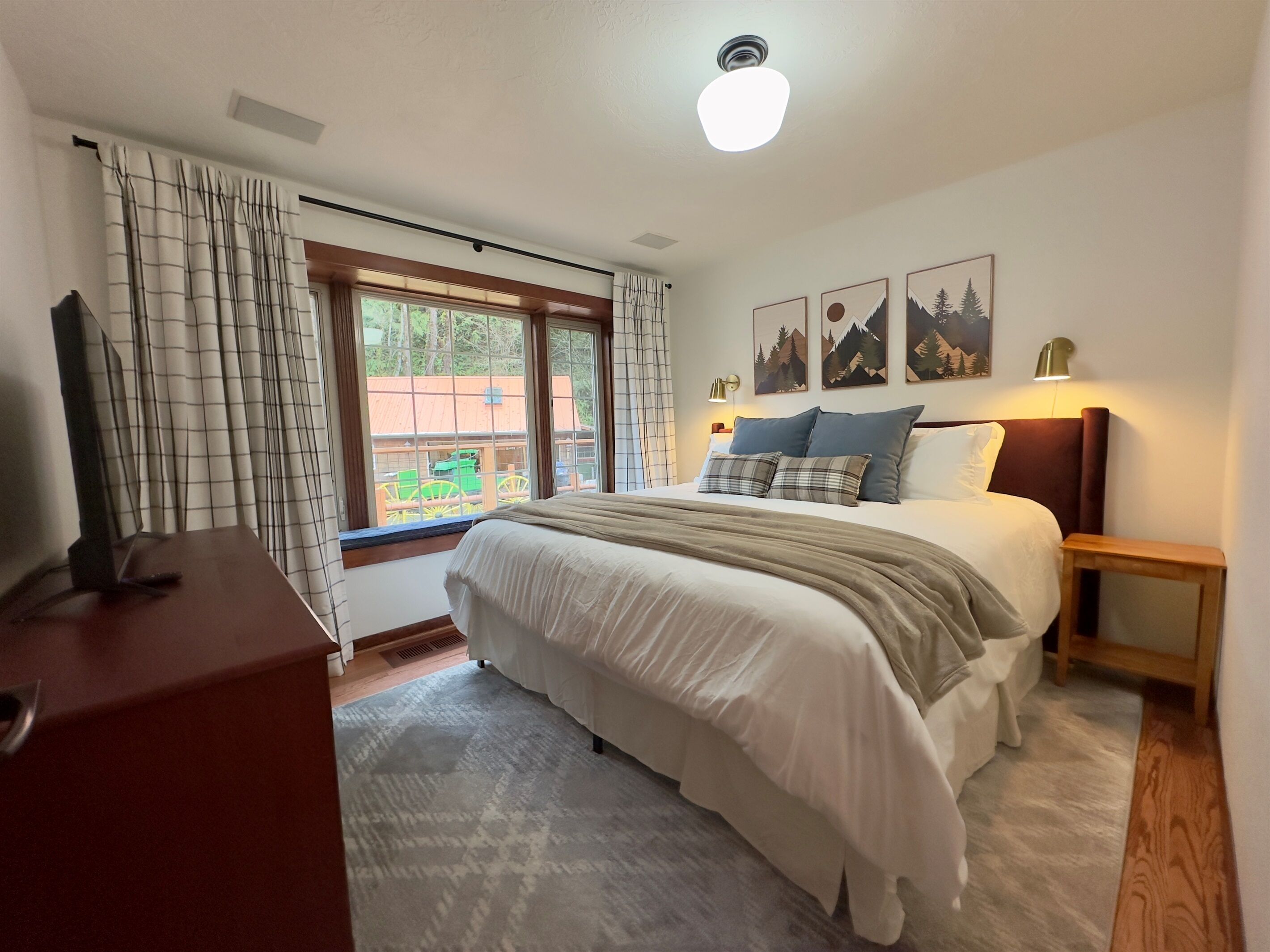 Een slaapkamer met een groot bed, een televisie op een houten dressoir, een raam met gordijnen en kunst aan de muur.
