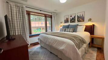 Een slaapkamer met een groot bed, een televisie op een houten dressoir, een raam met gordijnen en kunst aan de muur.