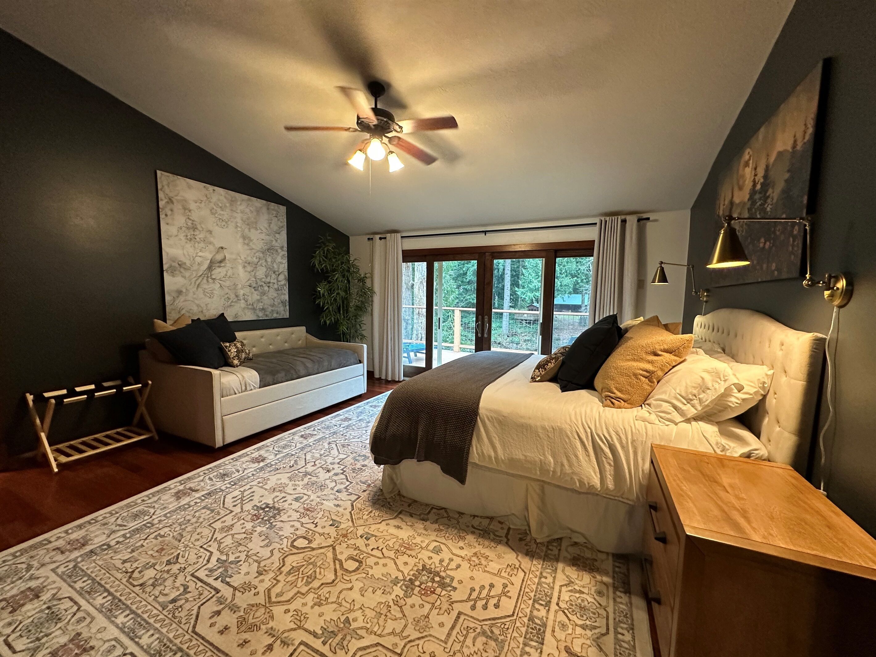 A bedroom with a bed, a sofa, a ceiling fan, a large window, and a view of the outdoors.