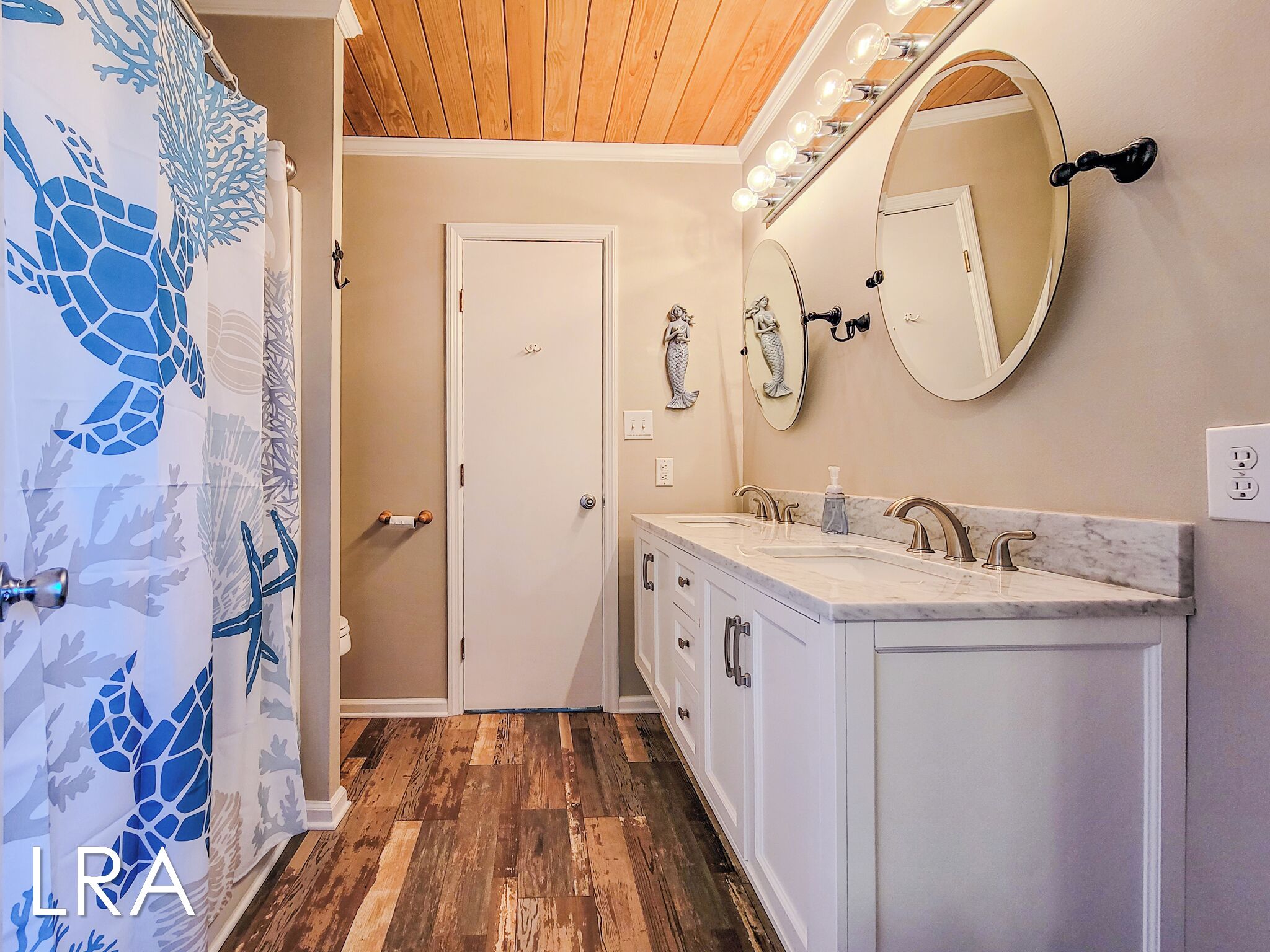 A bathroom with a wooden ceiling, a white door, a vanity with a mirror, and a shower with a blue sea turtle design.