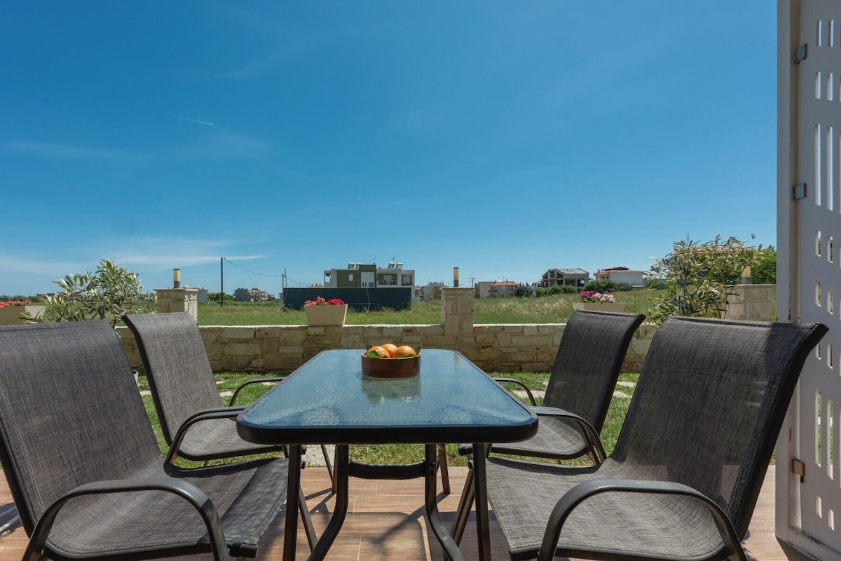 An outdoor patio with a table and four chairs, a bowl of fruit on the table, and a view of houses in the background.
