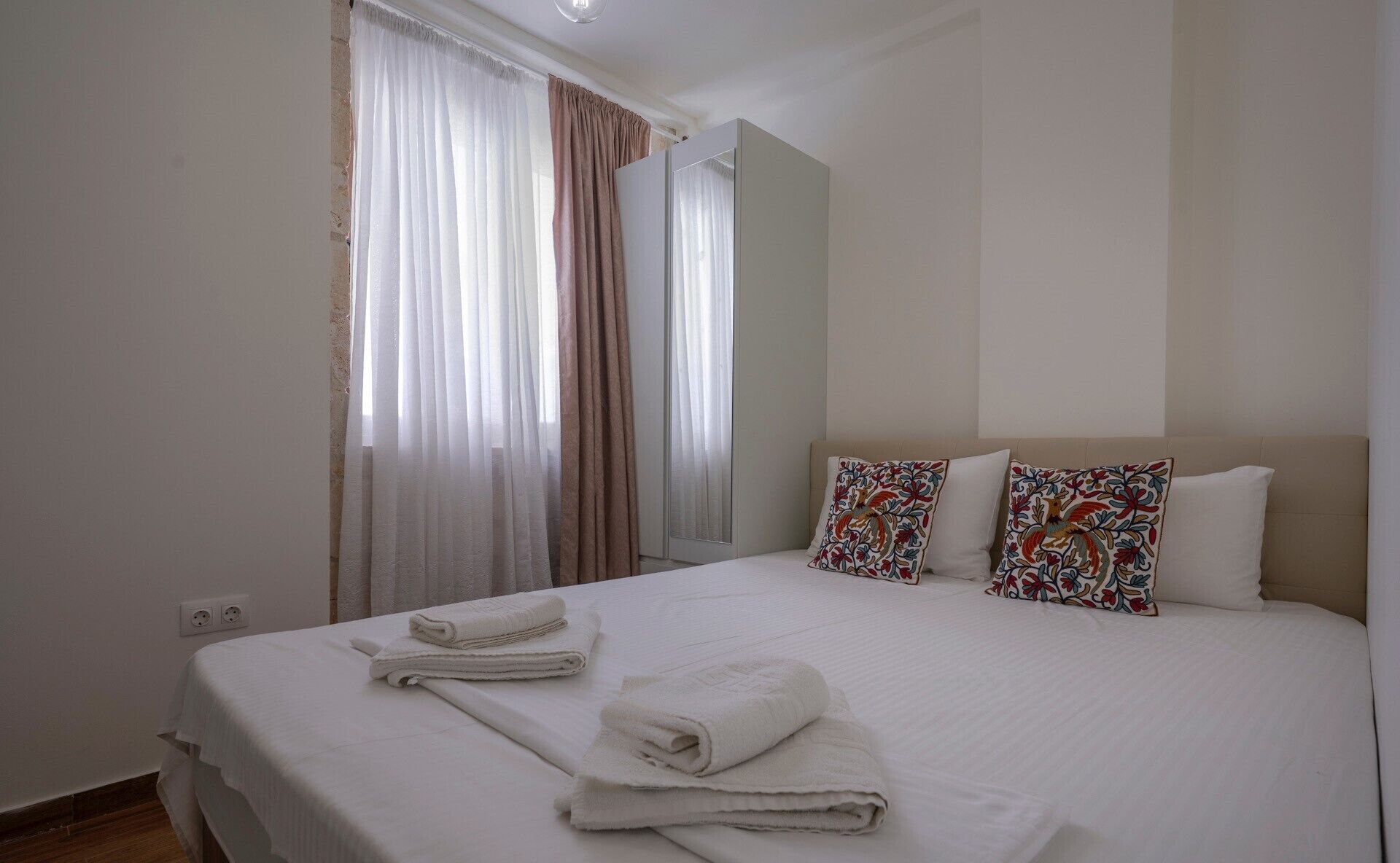 A hotel room with a large bed, two decorative pillows, a window with sheer curtains, and a mirror.