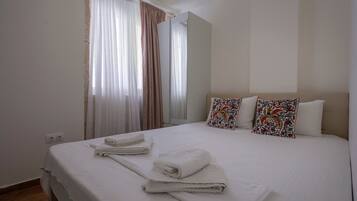 Una habitación de hotel con una cama grande, dos almohadones decorativos, una ventana con cortinas traslúcidas y un espejo.