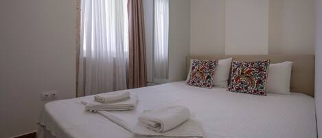 A hotel room with a large bed, two decorative pillows, a window with sheer curtains, and a mirror.