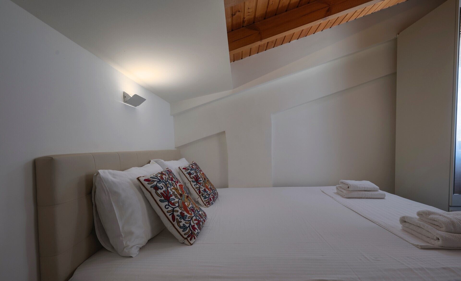 Un dormitorio con una cama grande, ropa de cama blanca y almohadas decorativas. Se ve una ventana al lado derecho y un techo de madera.