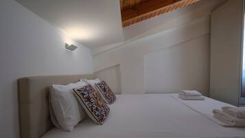 Un dormitorio con una cama grande, ropa de cama blanca y almohadas decorativas. Se ve una ventana al lado derecho y un techo de madera.