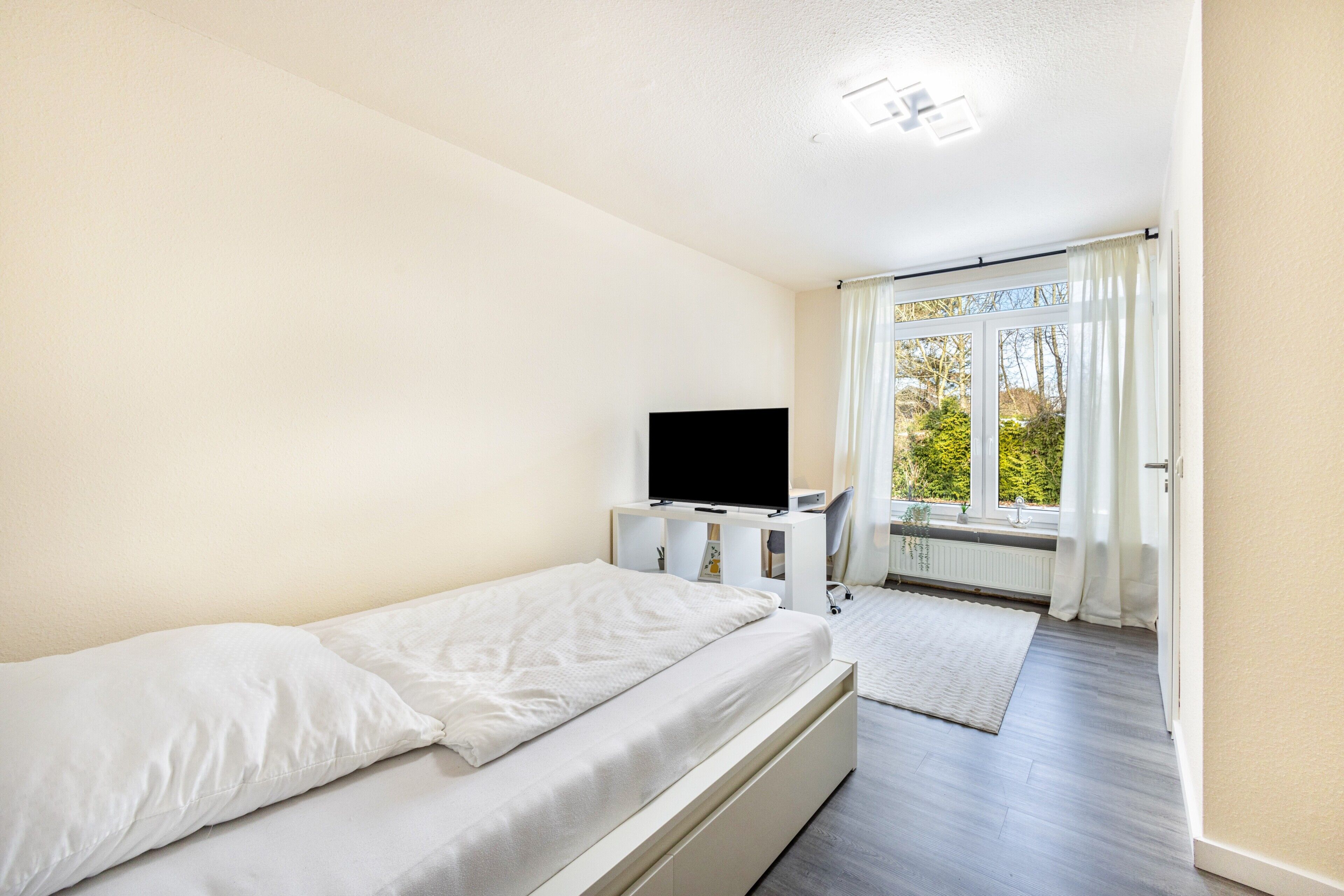 Une chambre à coucher avec un lit, un téléviseur posé sur un meuble, une fenêtre avec des rideaux et une vue sur la verdure à l’extérieur.