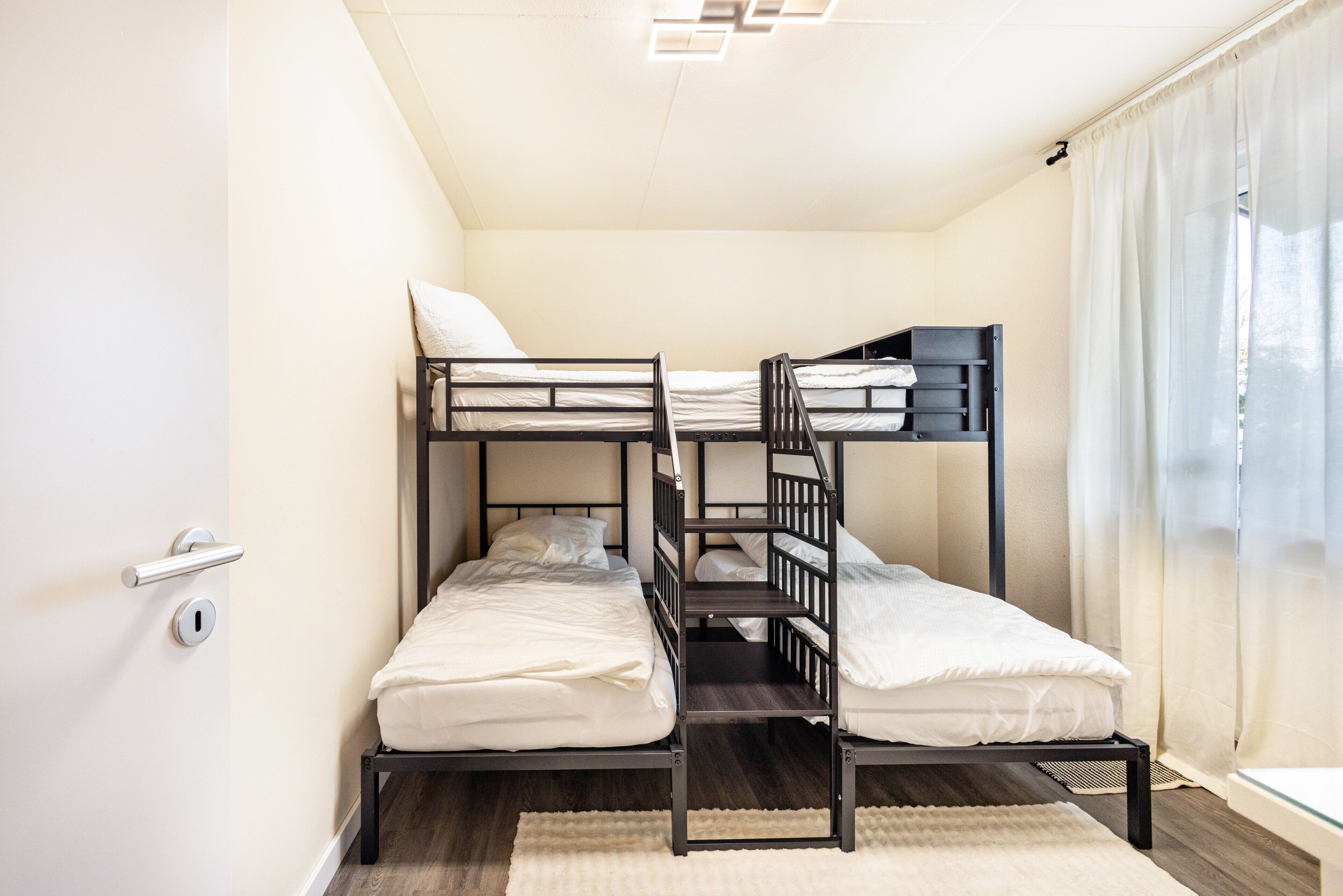Habitación con literas, una puerta, una ventana con cortinas y una lámpara en el techo.
