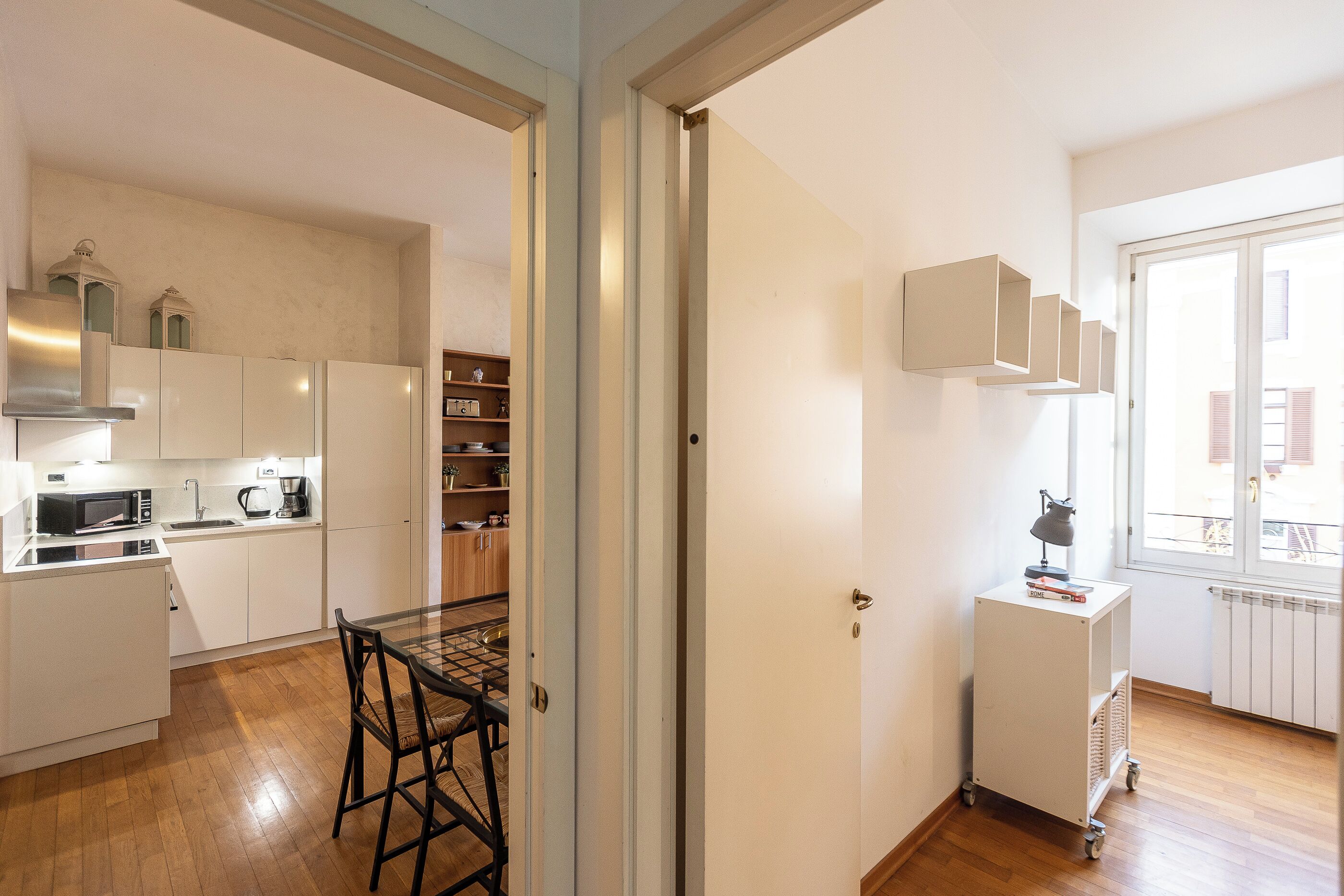 Eine moderne Küche mit weißen Schränken, einer Mikrowelle und einer Kaffeemaschine. Ein Essbereich mit Tisch und Stühlen ist vorhanden. Im Hintergrund ist ein weißer Schrank mit Regalen zu sehen.