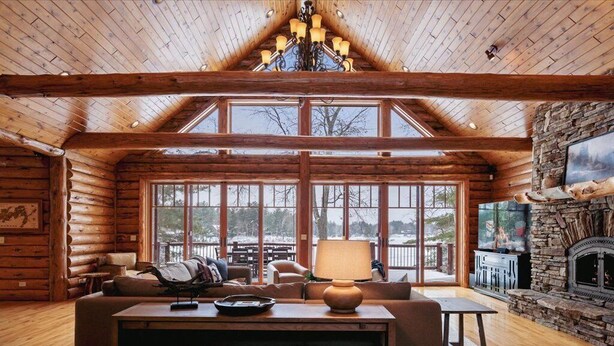 Ein Holzblockhaus-Interieur mit großem Fenster, Kamin und Holzbalkendecke.