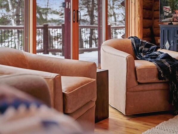 A cozy living room with a brown leather sofa, a wooden coffee table, and a view of a wooden deck through large glass doors. - WFC Luxury Cabin Daggett Lake|Beach|Kayaks|Firepit (Crosslake)