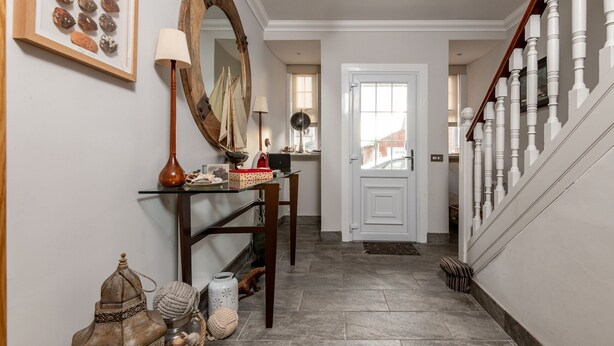 A hallway with a wooden staircase, a mirror, a framed picture of rocks, a lamp, and a small table with decorative items. - Main Street (No 115) | Lower Largo (Lower Largo)