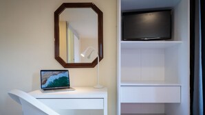 Une chambre moderne avec un bureau blanc, un ordinateur portable, un miroir, un téléviseur posé sur une étagère et une chaise blanche.