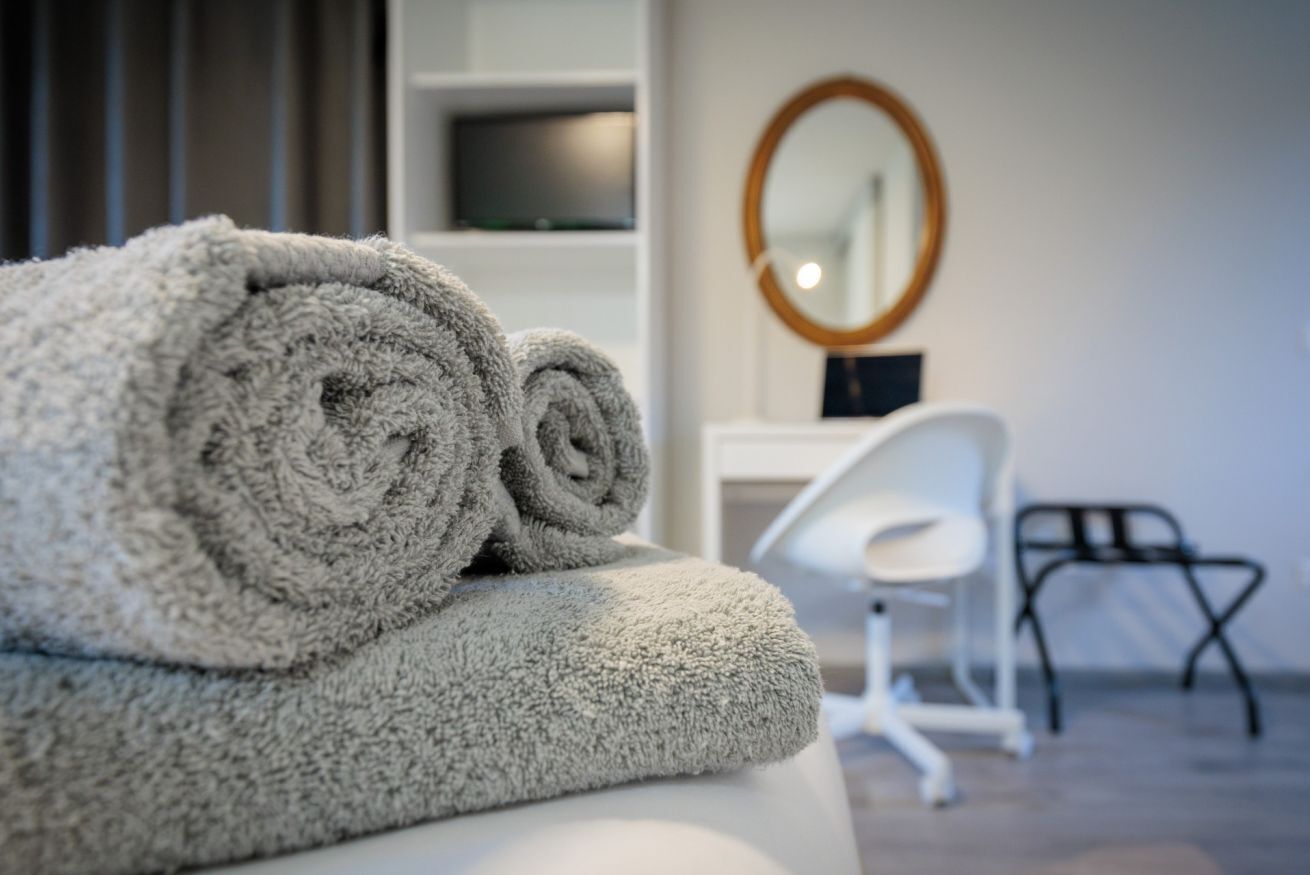 Une serviette soigneusement pliée sur une surface blanche, une chaise blanche et un miroir dans une pièce moderne, avec un téléviseur posé sur une étagère.