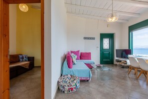 A room with a bed, a sofa, a dining table with chairs, a TV, and a view of the sea.