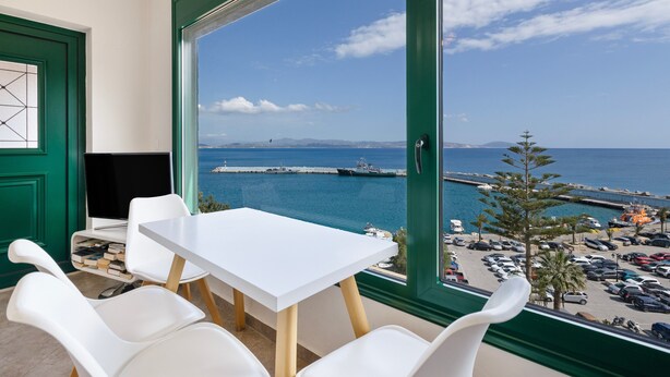 A room with a large window offering a view of a harbor, a table, and white chairs. - Apartment 'Two Bedroom  Sea View' with Sea View, Wi-Fi and Air Conditioning (Crete)