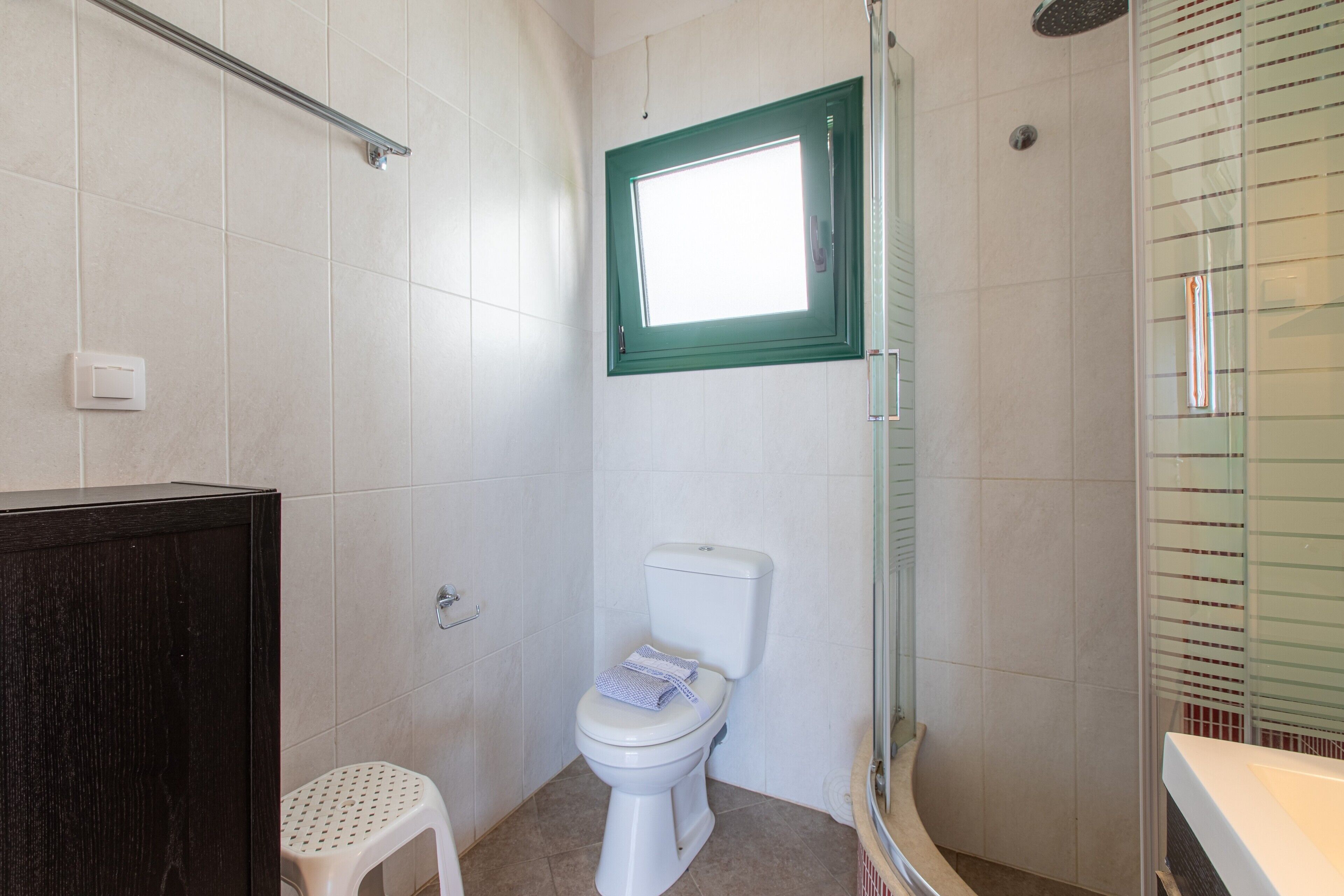Un baño con inodoro blanco, un taburete pequeño, cabina de ducha de vidrio y ventana con marco verde.