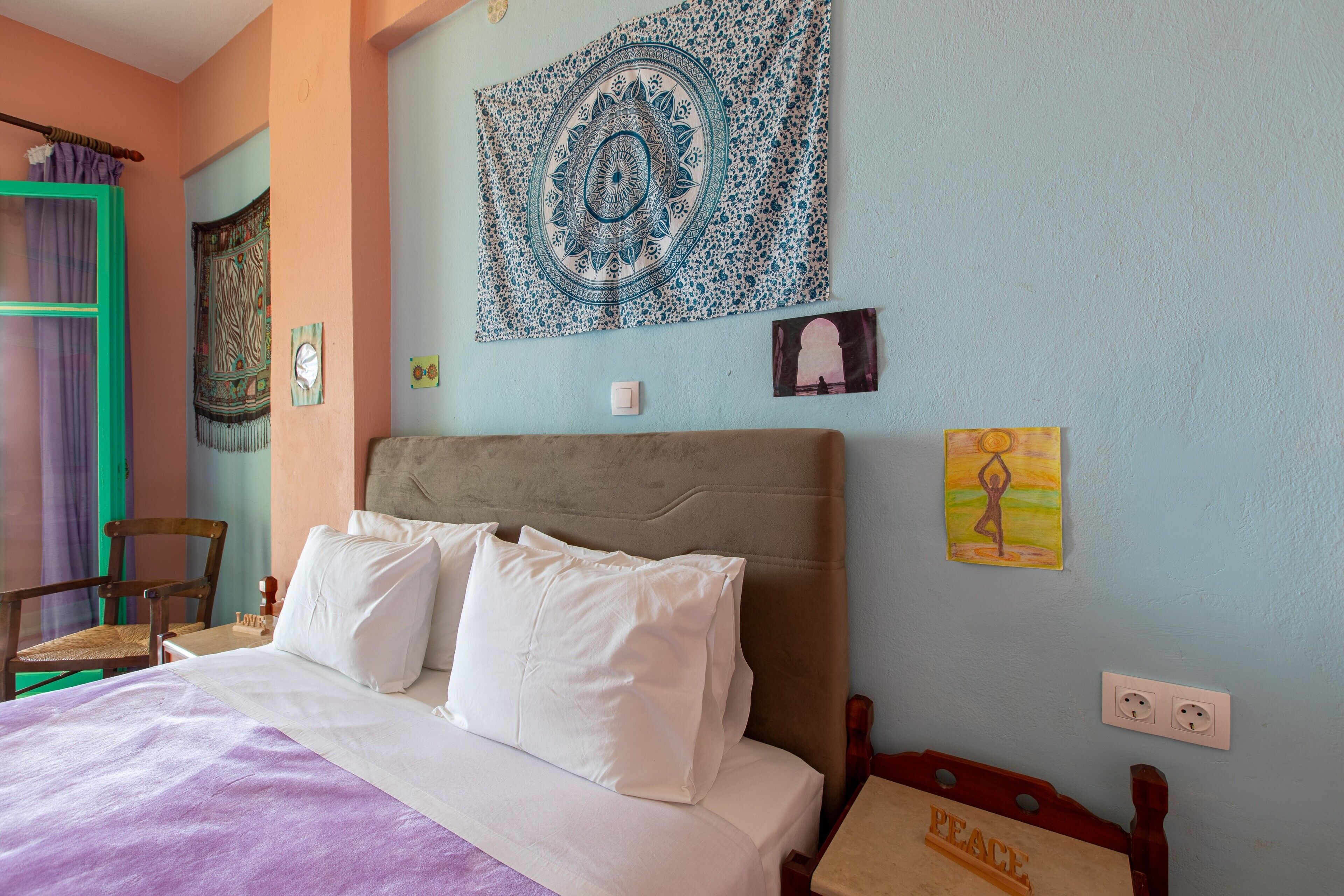 Una cama con ropa de cama blanca, cabecero y pared decorada con un tapiz y obras de arte.