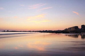Ein Strand mit ruhigem Wasser, das den Himmel spiegelt, ein ferner Stadtpanorama und einige Gebäude entlang des Ufers.