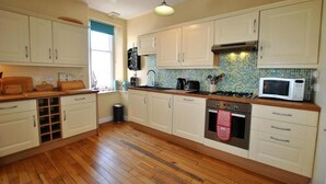 A kitchen with wooden flooring, white cabinets, a microwave, a stove, and a window with blue curtains. - Bayview Townhouse | St Monans (St Monans)