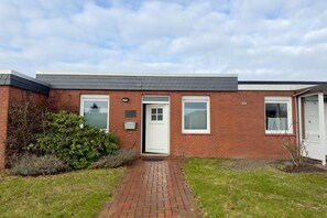 A brick building with a white door, windows with blinds, and a small garden area with a brick pathway. - Bungalow mit Garten in Dornumersiel (Dornumersiel)