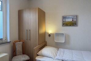 A bedroom with a bed, a wooden wardrobe, a chair, a wall-mounted shelf, and a framed picture of a lighthouse. - Bungalow mit Garten in Dornumersiel (Dornumersiel)