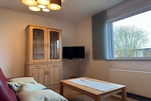 A room with a wooden cabinet, a television, a window, and a table. - Bungalow mit Garten in Dornumersiel (Dornumersiel)