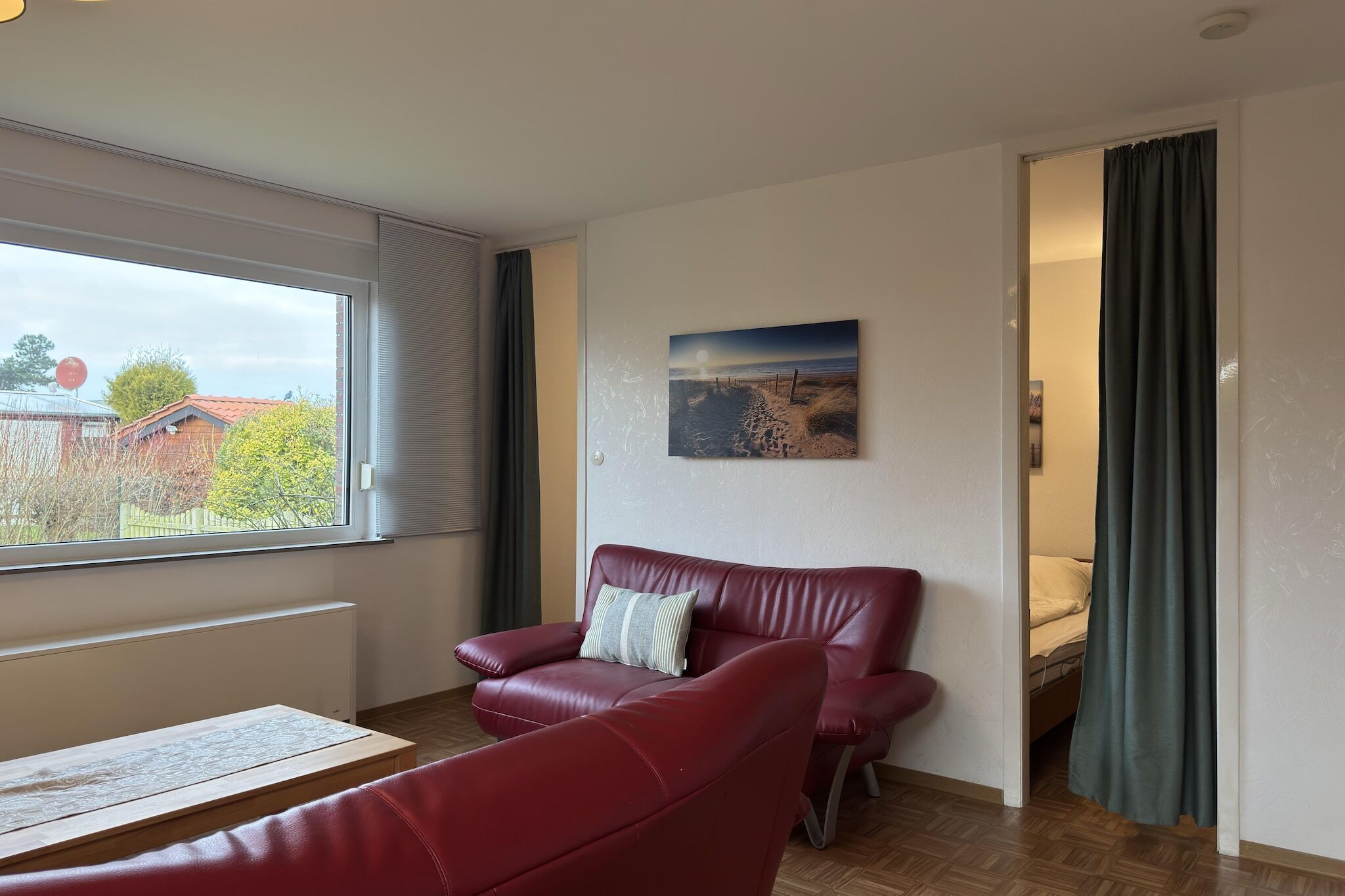 Een kamer met een rode leren bank, een houten tafel, een schilderij aan de muur en uitzicht door een raam.